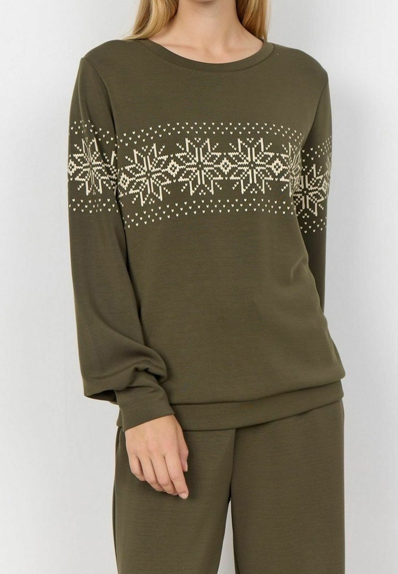Femme portant un haut à manches longues vert olive et un pantalon avec des motifs de flocons de neige de couleur crème sur la poitrine et le haut des manches.