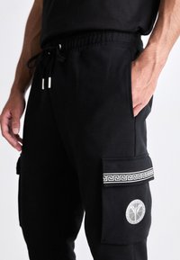 Zwarte joggingbroek met een trekkoord in de taille, zijzakken, een decoratieve zilveren band en een embleem op de cargozak. Zachte stoffen textuur.