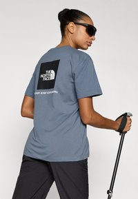 Γκρι βαμβακερό t-shirt με μαύρο τετράγωνο σχέδιο που γράφει "THE NORTH FACE" και "NEVER STOP EXPLORING", συνδυασμένο με σκούρο παντελόνι και μπαστούνι πεζοπορίας.