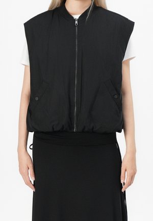 Kvinde iført en svart, ærmeløs vest med lynlås over en hvid kortærmet skjorte og et sort skørt, stående mod en hvid baggrund.