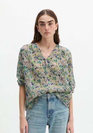 Frau mit langen braunen Haaren trägt eine lockere, mit Blumen gemusterte Bluse mit aufgerollten Ärmeln und hellblauen, hoch taillierten Jeans vor weißem Hintergrund.