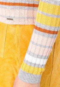 Mehrfarbiger gestreifter Strickpullover in Rosa, Orange, Grau und Gelb. Gerippte Textur mit grauem Bund. Kombiniert mit einer gelben Wildlederhose.