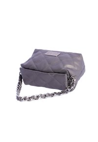 Bolso de mano de cuero gris acolchado con una correa de cadena plateada y una placa metálica con el logo. Presenta una forma estructurada y una textura suave.