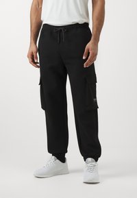 Mann trägt schwarze Cargohosen mit Kordelzug am Bund und weiße Sneakers, steht vor einem schlichten weißen Hintergrund.