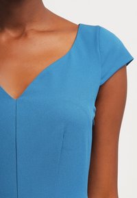 Frau in einem kurzärmligen blauen Kleid mit einem breiten V-Ausschnitt, der einen Teil des Halses und des oberen Brustbereichs zeigt.