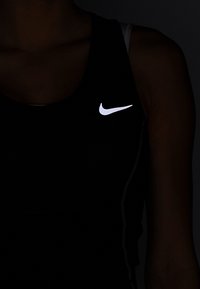 Débardeur de sport noir avec texture en maille, arborant un logo Nike réfléchissant blanc sur la poitrine et de fines coutures blanches sur les côtés.