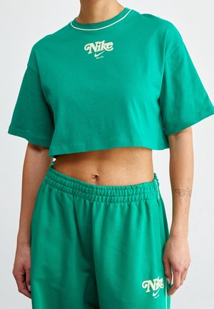 Person trägt grünes Nike-Bauchfrei-Top und passende Shorts mit elastischem Bund, zeigt Bauch und Schmetterlingstattoo am linken Unterarm.