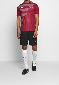 Punainen urheilupaita, jossa on valkoisia täpläkuvioita, mustat verkkarit, valkoiset sukat ja mustat nappulakengät; vaatteissa ja kengissä on Puma-logo.