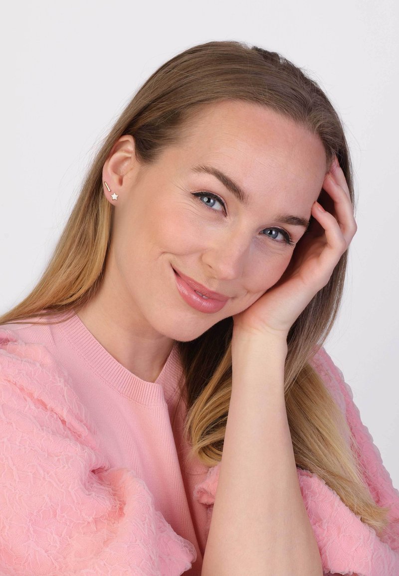 Des boucles d'oreilles en forme d'étoile dorée et de ligne portées par une femme aux cheveux longs. Elle porte un pull texturé rose clair et se trouve devant un fond neutre.