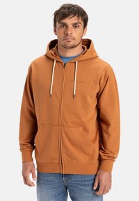 Brun zip-up hoodie med dragsko på huvan, med två framfickor och en texturerad tyg. Bärs över en blå skjorta.