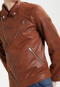 Giacca in ecopelle marrone con colletto, chiusure a zip e dettagli a coste sulle spalle. Texture liscia, design aderente e silhouette angolare.