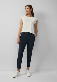 Haut sans manches blanc avec un design imprimé texturé, associé à un pantalon ajusté bleu marine et des baskets blanches. Tissu lisse et coupe ajustée.