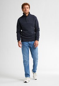 Bărbat cu păr creț, îmbrăcat într-un pulover bleumarin, blugi albaștri și adidași albi, mergând pe un fundal alb simplu.