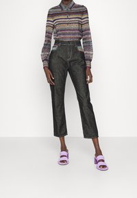 Kvinna klädd i en flerfärgad zigzag långärmad skjorta, högmidjade svarta jeans med färgglada fickdetaljer och lavendelfärgade klacksandaler.