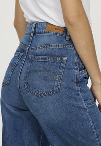 Jeans en denim bleu taille haute avec une coupe décontractée, dotés de coutures orange, de deux poches arrière et d'un patch en cuir marron.