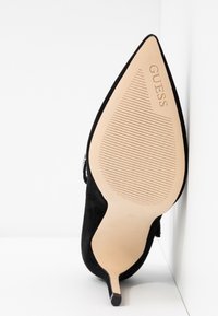 Chaussure noire à talon haut pointu, inclinée vers le haut, montrant la semelle beige avec le logo "GUESS" et des lignes antidérapantes texturées.