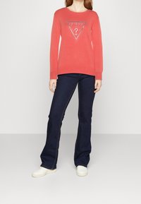 Maglia a maniche lunghe color corallo con logo triangolare metallico Guess, abbinata a jeans svasati blu scuro e sneakers bianche. Outfit semplice e casual.