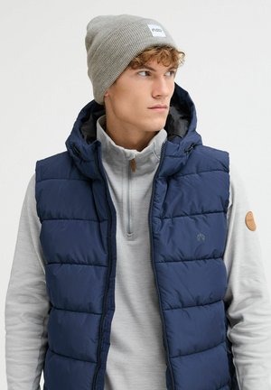 Marineblå puffer vest med hette, med horisontal quilt. Brukt over en lys grå zippered topp, complementert av en lys grå lue.
