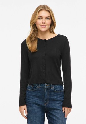 Vrouw die een zwart, met knopen gesloten, top met lange mouwen en een blauwe spijkerbroek draagt, staand tegen een effen witte achtergrond.