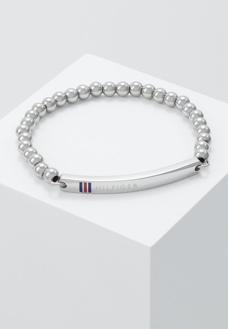 Bracelet perlé argenté avec une plaque métallique incurvée gravée de "HILFIGER" et un petit logo de drapeau rouge, blanc et bleu.