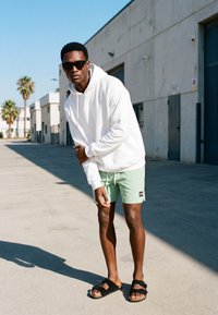 Ung mand iført solbriller, hvid hættetrøje, mintfarvede shorts og sorte sandaler, står og lener sig fremad på en solrig byfortov.