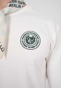 Camisa blanca con un logo circular verde oscuro de "Prohibited Motorsport Company Berlin" en el pecho y una bufanda ligera azul clara con estampado alrededor del cuello.