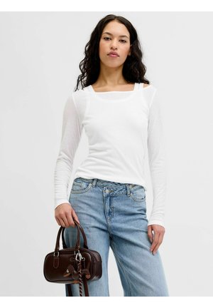 Mladá žena s dlhými tmavými vlasmi má na sebe biele tričko s dlhým rukávom a modré džínsy, drží malú hnedú kabelku so zvončekom v tvare kačky.