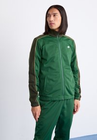 Ensemble de survêtement vert avec une veste zippée et des poignets élastiques, comportant un logo blanc et des accents dorés le long des coutures, fabriqué en tissu lisse.
