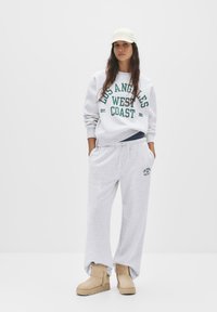 Grijze sweatshirt met de opdruk "Los Angeles West Coast", gecombineerd met grijze trainingsbroek en tan laarzen; een pet maakt de casual look af.