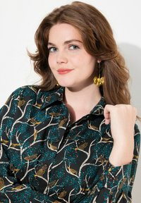 Camicia nera con un motivo di uccelli e rami in oro e teal. Presenta un colletto a bottoni e maniche lunghe, abbinata a orecchini floreali gialli.