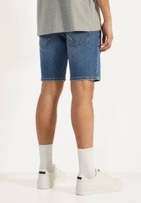 Man i knälånga blå denimshorts, vita räfflade strumpor, vita sneakers och en grå texturerad skjorta, stående på ett vitt golv.