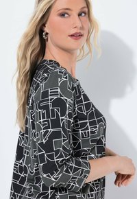 Musta ja harmaa toppi, jossa on abstrakteja valkoisia geometrisia kuvioita, kolmannen neljänneksen hihat ja pyöreä kaula-aukko. Sujuva kankaankuvio ja rento istuvuus.