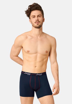 Uomo senza maglietta con barba corta che indossa boxer blu navy con cuciture rosse e fascia in vita con scritta "sloggi", in piedi contro uno sfondo semplice.