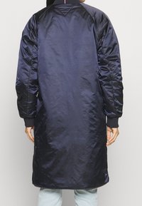 Lang marineblå bomberjakke med shiny satinfinish, ribbet krave og tætsiddende manchetter. Har sidelommer og en slank, strømlinet design.