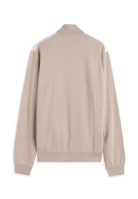 Chaqueta beige con cremallera, presenta un cuello alto y rayas blancas en los hombros. Fabricada con tejido liso, con mangas largas y puños acanalados.