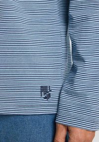 Chemise à rayures bleues et blanches à manches longues en tissu texturé. Présente un petit logo brodé en bas. Design décontracté.