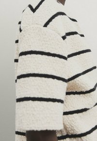 Sweter robiony na drutach w kremowym kolorze z czarnymi poziomymi paskami. Posiada teksturowany materiał i krótkie rękawy. Wzór zawiera dekolt w łódkę.