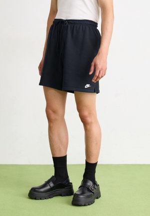 Osoba nosi czarne szorty Nike z troczkiem, czarne skarpetki i czarne masywne buty z klamrą, stoi na zielonej podłodze na tle białej ściany.