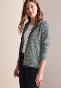Veste zippée à capuche en tissu vert texturé avec un motif en chevron, ornée d'une bordure bleu marine et de deux poches avant.
