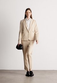 Béžový textúrovaný oblek s upraveným sakom a zodpovedajúcimi nohavicami, kombinovaný s bielou košeľou a malou čiernou kabelkou. Čierne mokasíny dotvárajú celé oblečenie.