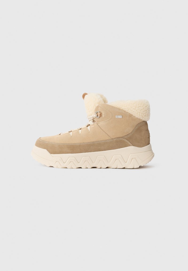 Botas de tobillo de ante en color marrón con un cuello de crema esponjoso, con una suela gruesa de patrón en zigzag y cordones. Acentos metálicos en el lateral.
