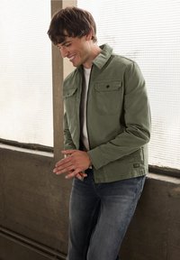 Kurtka w kolorze oliwkowej zieleni z zamkiem z przodu, dwoma kieszeniami na piersi i stójką, zestawiona z niebieskimi dżinsami w swobodnym otoczeniu.