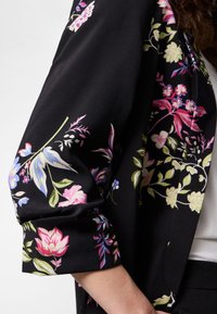 Black fabric with colorful floral patterns in pink, purple, green, and blue draped over a bent arm against a white background.