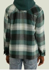 En person som bär en grön, svart och vit rutig jacka med grön huva, kombinerad med ljusblå jeans, vänd bort från kameran.