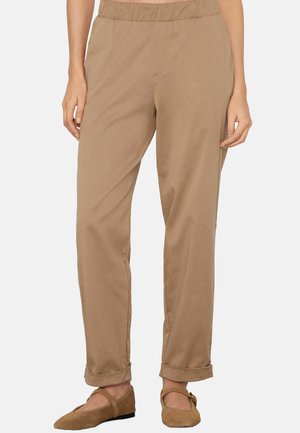 Donna che indossa pantaloni beige a gamba stretta con orli arrotolati e ballerine Mary Jane marroni abbinate, in piedi con le mani rilassate ai lati.