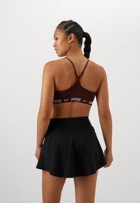 Bordó sportmelltartó fehér Nike logóval és keresztben futó pántokkal, fekete, bővülő sportszoknyával párosítva, sima textúrával.