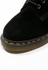 Botín de ante negro hasta el tobillo con punta redondeada, que presenta una suela texturizada y una costura amarilla a lo largo del borde. Los cordones son negros.