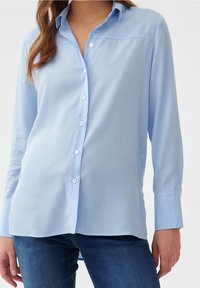 Camisa de botones azul claro con una textura suave, mangas largas y un collar clásico. Cuenta con un dobladillo recto y un sutil yugo en los hombros.