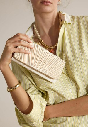 Vrouw in een geel gestreept shirt houdt een geplooide beige clutch vast met een gouden armband en gelaagde kettingen met bedels.