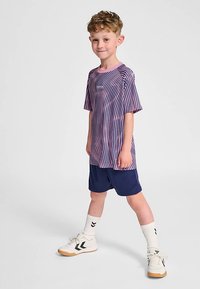 Gestreiftes T-Shirt mit kurzen Ärmeln in Lila und Pink, kombiniert mit marineblauen Shorts. Weiße und schwarze Turnschuhe sowie weiße Socken mit schwarzem Akzent.